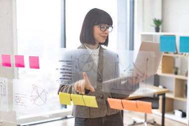 Modern ofisteki cam tahtadaki çizelgelerin büyümesini analiz ederken dijital dizüstü bilgisayar kullanan dalgın bir iş kadını. İş stratejisi ve dijital pazarlama kavramı.