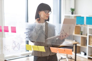 Modern ofisteki cam tahtadaki çizelgelerin büyümesini analiz ederken dijital dizüstü bilgisayar kullanan dalgın bir iş kadını. İş stratejisi ve dijital pazarlama kavramı.