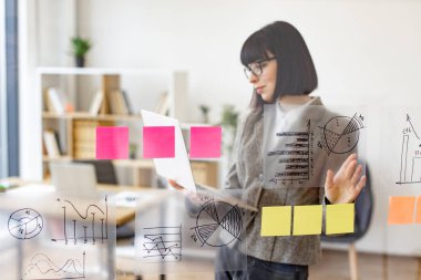 İşteki sunuma hazırlanan kadın yönetici. Gözlüklü beyaz kadın dijital laptop tutuyor ve ofis toplantısından önce cam duvardaki finansal grafiklere bakıyor..