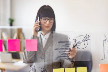 Modern ofisteki cam panodaki çizelgelerin büyümesini analiz ederken dalgın bir iş kadını telefonda konuşuyor. İş stratejisi ve dijital pazarlama kavramı.