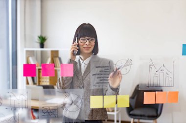 Modern ofisteki cam panodaki çizelgelerin büyümesini analiz ederken dalgın bir iş kadını telefonda konuşuyor. İş stratejisi ve dijital pazarlama kavramı.