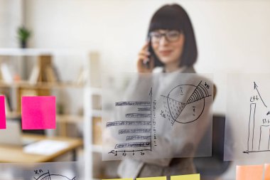 Gözlüklü ve şık takım elbiseli bayan ofis çalışanı dijital telefonu şirkette kullanıyor. Cam tahtanın önünde duran ve akıllı telefondan konuşan güzel bir genç kadının portresi..