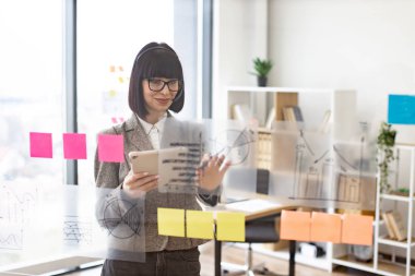 Ofiste video toplantısı için kulaklık ve tablet kullanan çekici bir iş kadınının ön görüntüsü. Cam duvarın yanında duran ve sohbet sırasında not alan odaklanmış genç bir kadın..