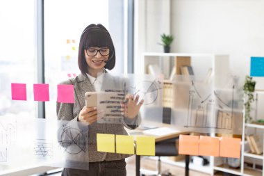 Ofiste video toplantısı için kulaklık ve tablet kullanan çekici bir iş kadınının ön görüntüsü. Cam duvarın yanında duran ve sohbet sırasında not alan odaklanmış genç bir kadın..