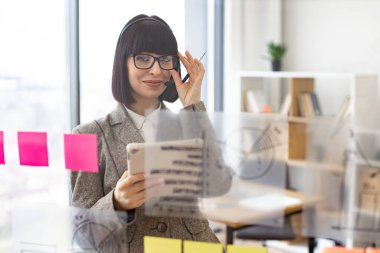 Elinde tablet olan akıllı iş kadını portresi yapışkan notlarla şeffaf cam duvara bakıyor. Gülümseyen kadın çalışan modern ofiste video konferansı yapıyor..