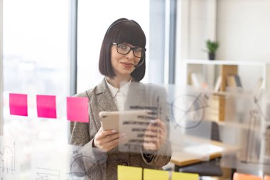 Elinde tablet olan akıllı iş kadını portresi yapışkan notlarla şeffaf cam duvara bakıyor. Gülümseyen kadın çalışan modern ofiste video konferansı yapıyor..