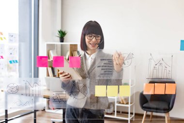 Toplantı odasında tabletten notlar alan genç ve ciddi bir bayan işçi. Konsantre olmuş, kulaklıklı beyaz kadın konferans salonundaki yapışkan kağıtlarla saydam tahtaya yazı yazıyor..