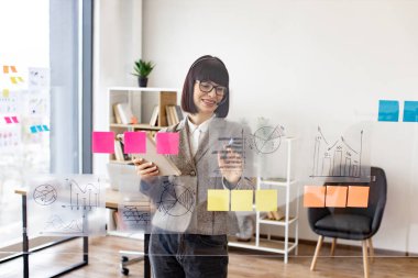 Toplantı odasında tabletten notlar alan genç ve ciddi bir bayan işçi. Konsantre olmuş, kulaklıklı beyaz kadın konferans salonundaki yapışkan kağıtlarla saydam tahtaya yazı yazıyor..