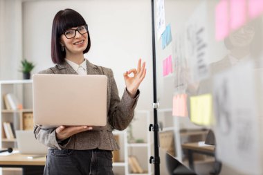 Gözlüklü ve şık takım elbiseli bir bayan ofis çalışanı dijital dizüstü bilgisayarla işaret veriyor. Cam tahtanın önünde duran ve kameraya gülümseyen güzel bir genç kadının portresi..