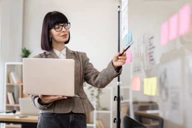 Sunuma hazırlanan kadın yönetici, gözlüklü genç kadın dizüstü bilgisayarlı çıkartmalar üzerine notlar yazıyor ve ofis toplantısından önce cam duvardaki finansal grafiğe bakıyor..