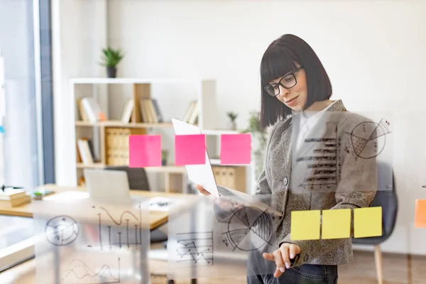 Female administrative manager preparing for presentation at work. Caucasian woman in glasses holding digital laptop and looking on financial graphic on glass wall before office meeting.