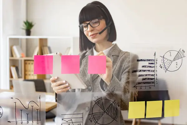 Caucasian woman holding tablet in hand while standing near transparent office board. Female entrepreneur in headset talking with business partner via video call and writing marks on sticky papers.
