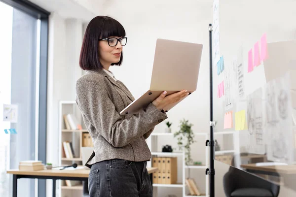 Confident caucasian woman in formal wear and glasses solving business issues with laptop in hands while standing at company boardroom. Concept of modern technology and busy people.