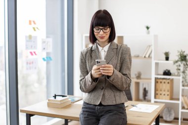 Gözlüklü neşeli beyaz iş adamı iş yerindeki cep telefonuyla dijital yazışmalara cevap veriyor. Takım elbiseli gülümseyen yönetici kablosuz bağlantı kullanarak elektronik iletişim geliştiriyor