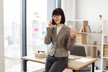 Gözlüklü neşeli beyaz kadın ve resmi kıyafetler masada otururken cep telefonuyla konuşuyor. Etkili girişimci iş konuşmalarını modern ofiste rahat bir şekilde yürütüyor..
