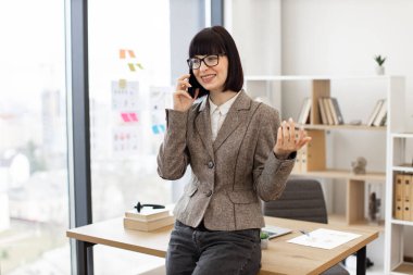 Gözlüklü neşeli beyaz kadın ve resmi kıyafetler masada otururken cep telefonuyla konuşuyor. Etkili girişimci iş konuşmalarını modern ofiste rahat bir şekilde yürütüyor..