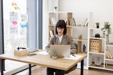 Bilgisayar görüntülerini incelerken proje detaylarını düşünen dikkatli bir iş adamı. Takım elbiseli ve gözlüklü genç beyaz kadın modern ofiste dizüstü bilgisayarla masada rahatça oturuyor..