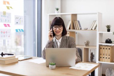 İş yerindeki dizüstü bilgisayarda web sayfalarını kaydırırken cep telefonuyla önemli aramalar yapan çekici beyaz bir insan. Ofisteki dijital cihazlar vasıtasıyla takım elbise pazarlığı yapan mutlu girişimci