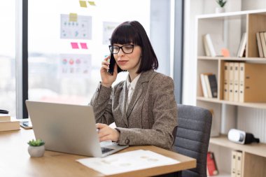 İş yerindeki dizüstü bilgisayarda web sayfalarını kaydırırken cep telefonuyla önemli aramalar yapan çekici beyaz bir insan. Ofisteki dijital cihazlar vasıtasıyla takım elbise pazarlığı yapan mutlu girişimci