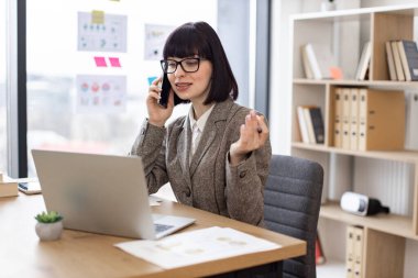Resmi giyinen neşeli bayan dijital ekrandan rapor okurken akıllı telefondan konuşuyor. Deneyimli çalışanlar iş yerlerindeki şirket verimliliğini artırma fikrini ortaya attılar..