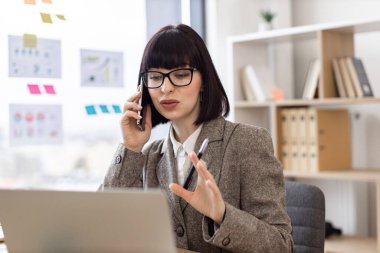 Takım elbiseli mutlu girişimci ofisteki dijital cihazlarla başlangıç alışverişi yapıyor. İş yerindeki dizüstü bilgisayarda web sayfalarını kaydırırken cep telefonuyla önemli bir arama yapan çekici beyaz kişi