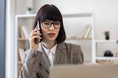 Ofisteki laptop ekranına bakarken cep telefonuyla konuşan gözlüklü çekici beyaz kadın. Verimli yönetici iş yerindeki müşteriyle iş görüşmesi sırasında bilgi paylaşımı yapıyor.