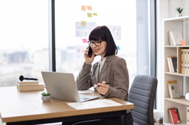 Verimli bir yönetici iş görüşmesi sırasında müşteriyle bilgi paylaşıyor. Ofisteki dizüstü bilgisayara bakarken cep telefonuyla konuşan gözlüklü çekici beyaz kadın..