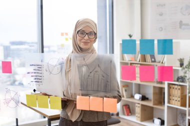 Cam tahtanın önünde duran ve kameraya gülümseyen müslüman genç bir kadının portresi. Şirkette dijital dizüstü bilgisayar kullanan tesettür ve şık takım elbiseli kadın ofis çalışanı..