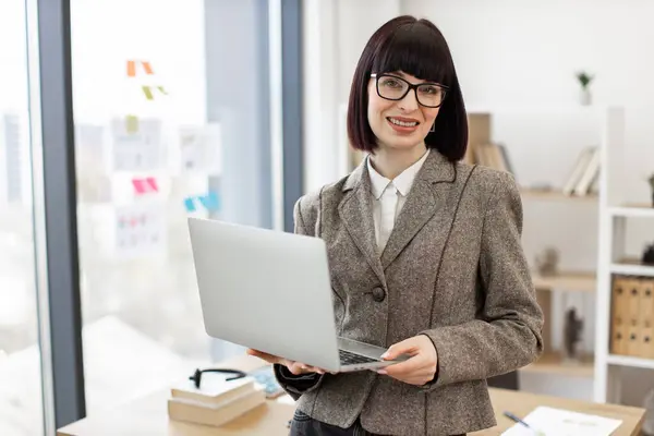 Cheerful woman in formal wear managing successful company from modern workplace. Portrait of smiling caucasian adult businesswoman sitting on edge of wooden desk with laptop.