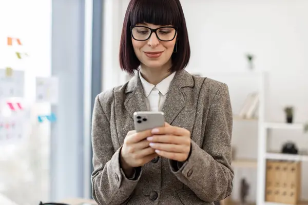 Joyful caucasian business person in glasses replying to digital correspondence via cell phone at work, copy space. Smiling manager in suit developing electronic communication using wireless connection