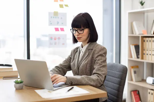 Focused caucasian woman in eyeglasses and formal wear working on laptop while sitting at office table. Professional entrepreneur searching for information over internet via technologies, copy space.