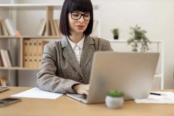 Young Caucasian lady in suit and eyeglasses sitting comfortably at table with laptop computer at modern office. Mindful business person thinking over project details while looking into display of pc.