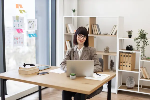 Mindful business person thinking over project details while looking into display of pc. Young Caucasian lady in suit and eyeglasses sitting comfortably at table with laptop computer at modern office.