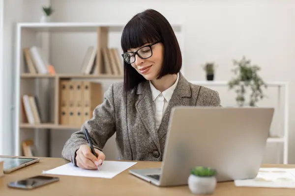 Mindful woman in eyeglasses making notes with pen while looking at laptop screen in workplace. Efficient Caucasian employee doing online research on companys development using wireless technologies.