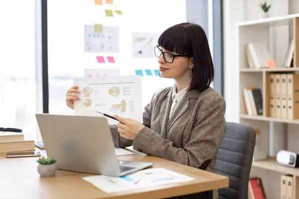 Efficient female manager showing sales report to colleagues in cozy workplace. Focused Caucasian woman in eyeglasses pointing at document with infographics while having online presentation via laptop.