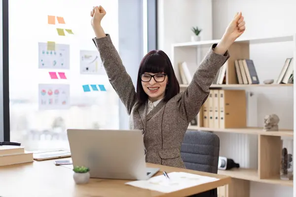 Emotional young lady in glasses holding hands high in air while screaming with happiness in modern office. Thrilled Caucasian specialist celebrating tender win with loud enthusiasm in workplace.