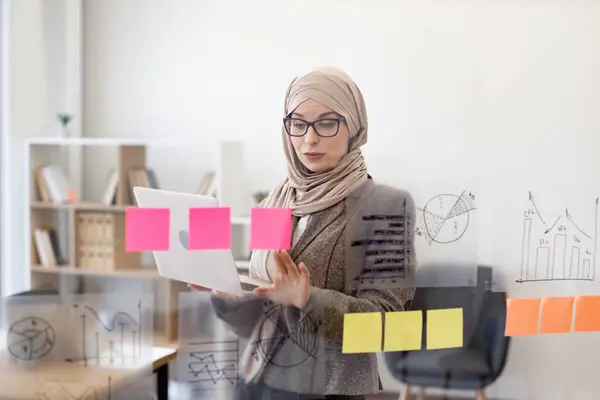 Confident muslim woman in formal wear and hijab solving business issues with laptop in hands while standing at company boardroom. Concept of modern technology and busy people.
