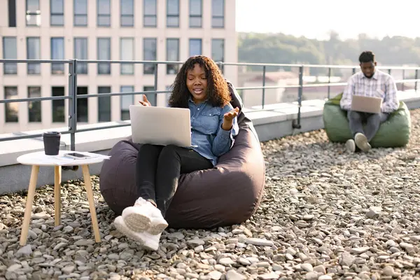 Happy African American female person in casual clothing gesture at laptop camera on flat roof of building, while her male colleague working in blurred background.