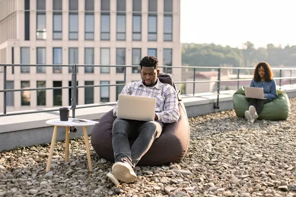 Successful business man dressed in checkered shirt and pants sitting at chair bag on roof top outdoors and typing on modern laptop, African American colleague works sitting in bag chair in background.