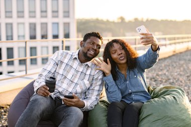 Genç Afrika kökenli Amerikalı kadın ve erkek öğle yemeği sırasında ofisin çatısında oturmuş selfie çekerken ya da cep telefonuyla video görüşmesi yaparken el sallıyorlar..