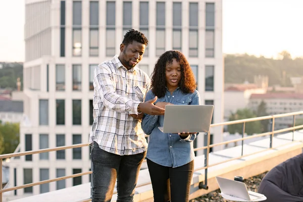Two African American business people outdoors during break standing on rooftop. Young adult man and woman using laptop while standing outdoors office building.