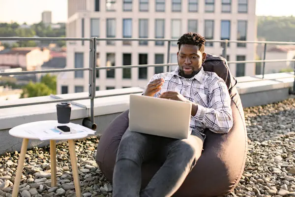Happy African American person in casual clothing gesture at laptop camera on flat roof of building. Efficient freelancer beginning video conference via internet connection outdoors.