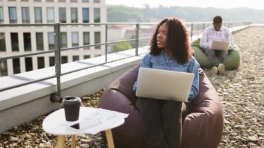 Çekici Afrikalı Amerikalı kadın serbest çalışan. Mobil bilgisayarı ofis çatısında uzaktan kumanda olarak kullanıyor. Mola sırasında koltukta oturuyor. İş arkadaşının bulanık geçmişi.