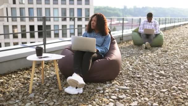 一位成功的商界女性穿着牛仔衬衫和裤子 坐在屋顶的椅袋前 在现代笔记本电脑上打字 而一位非洲裔美国同事则坐在椅背的椅背上工作 — 图库视频影像