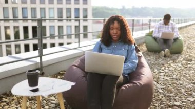 Açık havada dizüstü bilgisayar kullanan gülümseyen bir kadın, arka planda çalışan bir erkek arkadaş. Modern yaşam tarzı, bağlantı, iş dünyası, iletişim, web sohbeti, sosyal medya, video konferans kavramı.