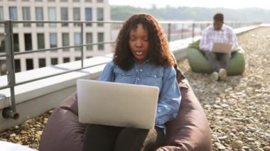 Açık havada dizüstü bilgisayar kullanan gülümseyen bir kadın, arka planda çalışan bir erkek arkadaş. Modern yaşam tarzı, bağlantı, iş dünyası, iletişim, web sohbeti, sosyal medya, video konferans kavramı.