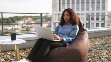 Sıradan kot gömlekli bir iş kadını dizüstü bilgisayarla çalışıyor. Profesyonel iş kadını dijital cihaz kullanıyor, görüntülü sohbet görüşmesi yapıyor, modern binanın çatısındaki armut çuvalında oturuyor..