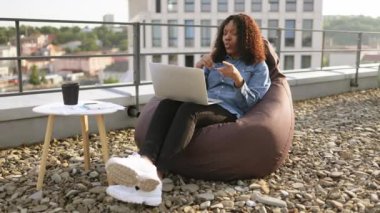 Düz çatısındaki laptop kamerasına günlük kıyafet jesti yapan mutlu Afrikalı Amerikalı. Açık havada internet bağlantısı üzerinden video konferansına başlayan etkili bir serbest yazar.