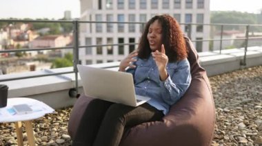 Düz çatısındaki laptop kamerasına günlük kıyafet jesti yapan mutlu Afrikalı Amerikalı. Açık havada internet bağlantısı üzerinden video konferansına başlayan etkili bir serbest yazar.