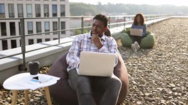 Başarılı bir iş adamı kot gömlek ve pantolon giyer açık çatıdaki koltuk çantasında oturur ve modern dizüstü bilgisayarda yazar, Afrikalı Amerikalı iş arkadaşı arka planda çanta koltuğunda oturur..